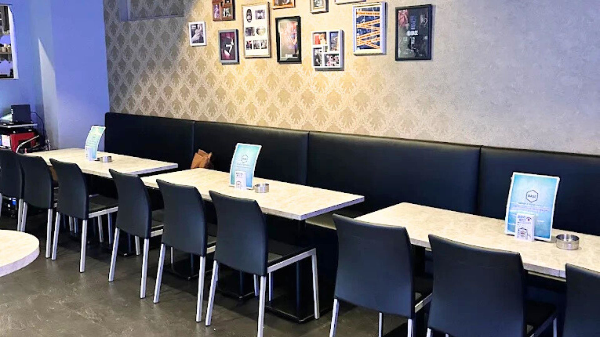 バーの座席エリアの別角度。長いソファ席と複数のテーブルが手前にあり、壁には装飾的な壁紙が貼られ、額縁に入った写真が飾られている。