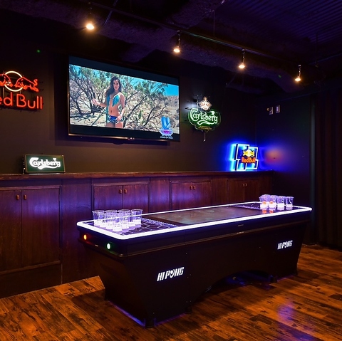暗いエンターテイメントルームに置かれた、LEDで光るビアポンテーブル。奥の壁にはテレビと、Red BullやCarlsbergのネオンサインが飾られている。