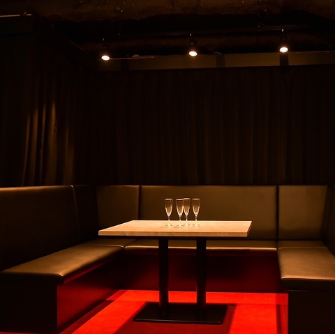 黒いL字型のソファに囲まれたテーブル。上には4つのシャンパングラスが置かれ、足元は赤い光で照らされているムーディーな空間。